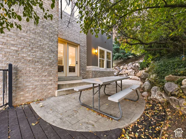 Patio area featuring light-toned masonry exterior, dual glass-paneled entry doors, composite deck, and natural stone retaining wall