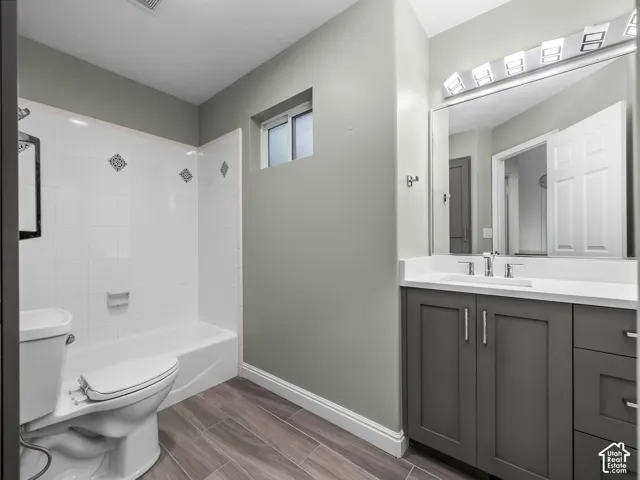 Bathroom featuring wood-finish flooring, a charcoal gray shaker-style vanity with white solid-surface countertop, a full-width vanity mirror with integrated multi-bulb lighting, and a white tile tub surround