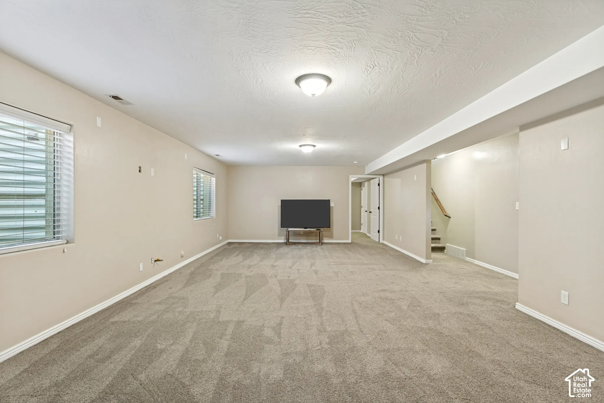 Neutral-toned carpeted living space featuring recessed lighting, dual windows with horizontal blinds, and a staircase with a wooden handrail