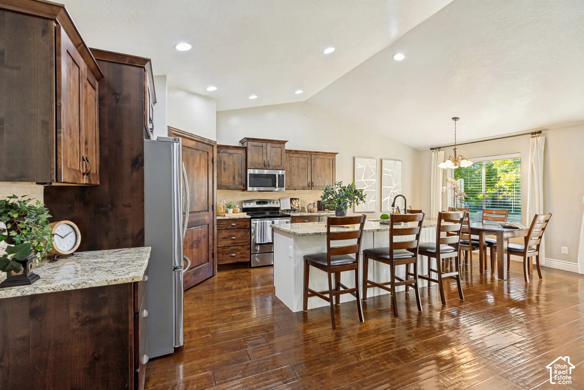 Kitchen featuring stained wood cabinetry, granite countertops, central island with bar seating, stainless steel appliances, and hardwood flooring under vaulted ceilings