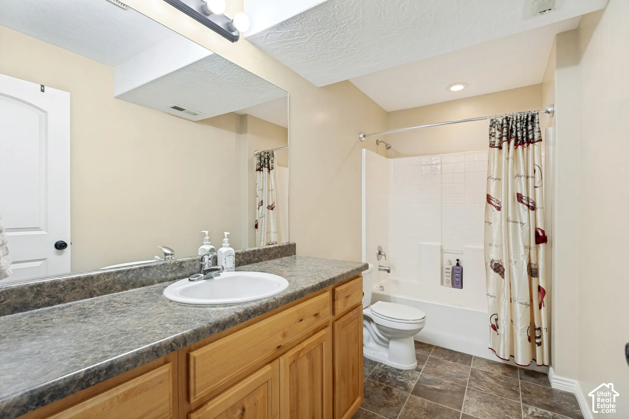 Bathroom featuring light wood-finish cabinetry, a neutral stone-pattern laminate countertop with a drop-in sink, a full-width vanity mirror, tile flooring, and a tub-shower combination with integrated wall shelving