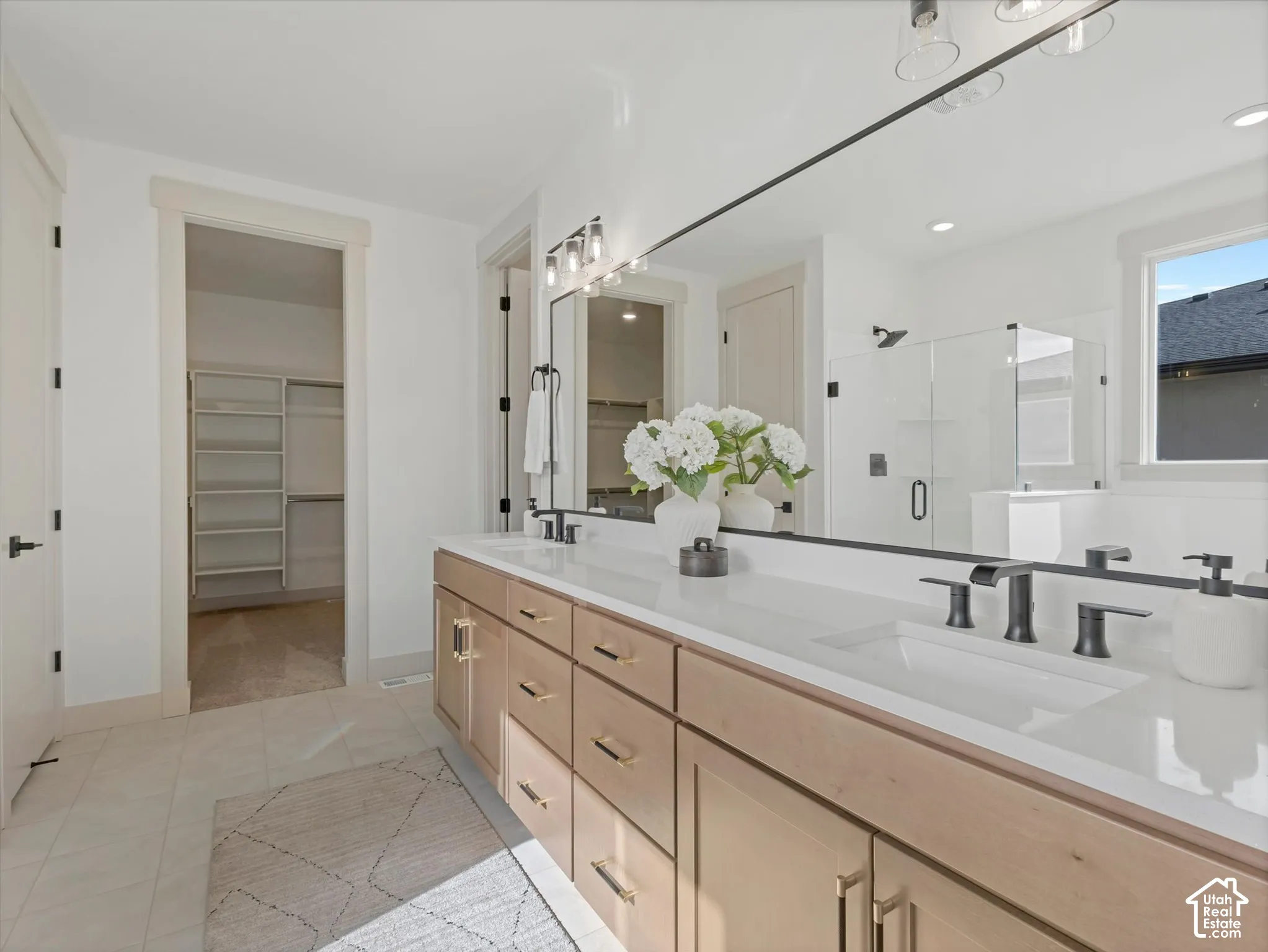 Double vanity with light-tone wood cabinetry, solid-surface countertop, rectangular undermount sinks, and matte black fixtures, featuring a full-width mirror and a walk-in closet with built-in shelving