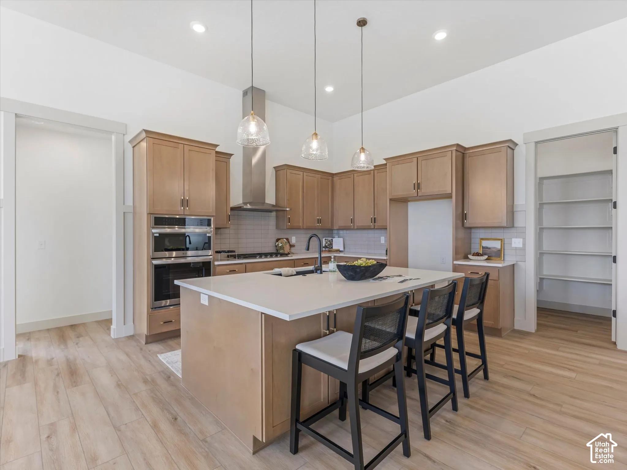 Kitchen featuring shaker-style cabinetry, quartz countertops, a central island with an integrated sink, built-in stainless steel double ovens, and wood-finish flooring