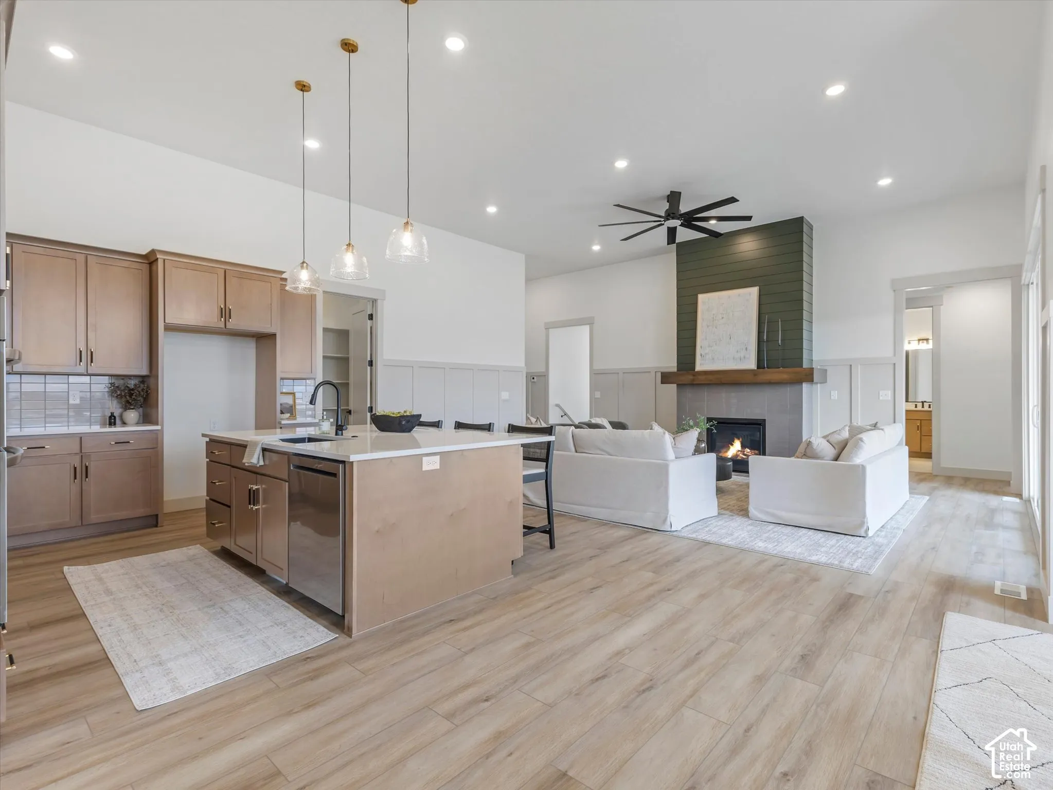 Open-concept kitchen and living area featuring wood-finish flooring, a central island with stone countertop, matte black hardware, three glass pendant lights, and a floor-to-ceiling fireplace with a wood mantel