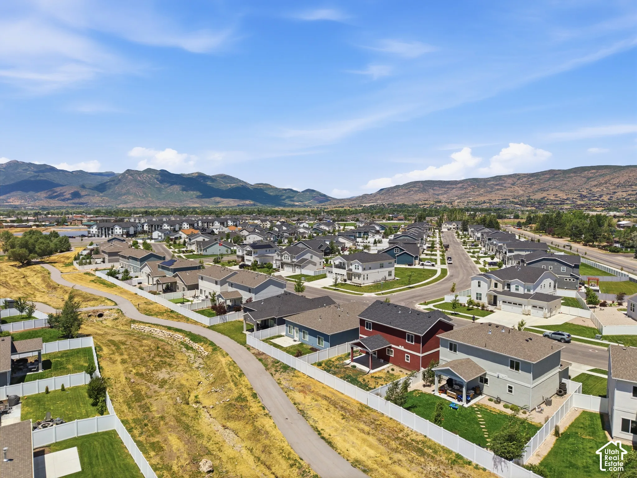 Aerial perspective of residential properties featuring paved walking trails, manicured lawns, white vinyl fencing, and mountain terrain in the distance