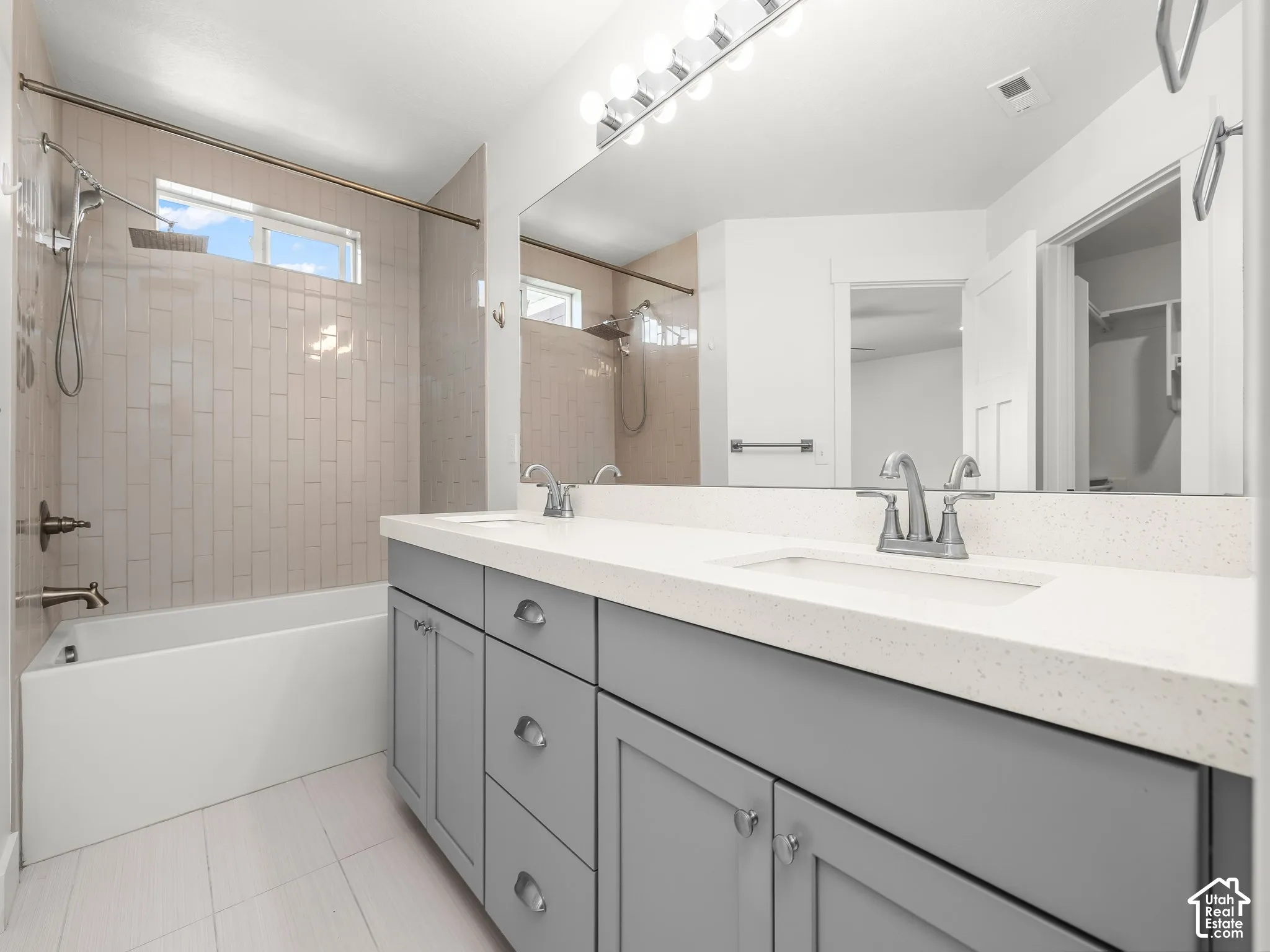 Dual-sink vanity with light-colored solid surface countertop and gray shaker-style cabinetry, full-width wall mirror, and tub-shower combination featuring vertically-laid tile wall surrounds