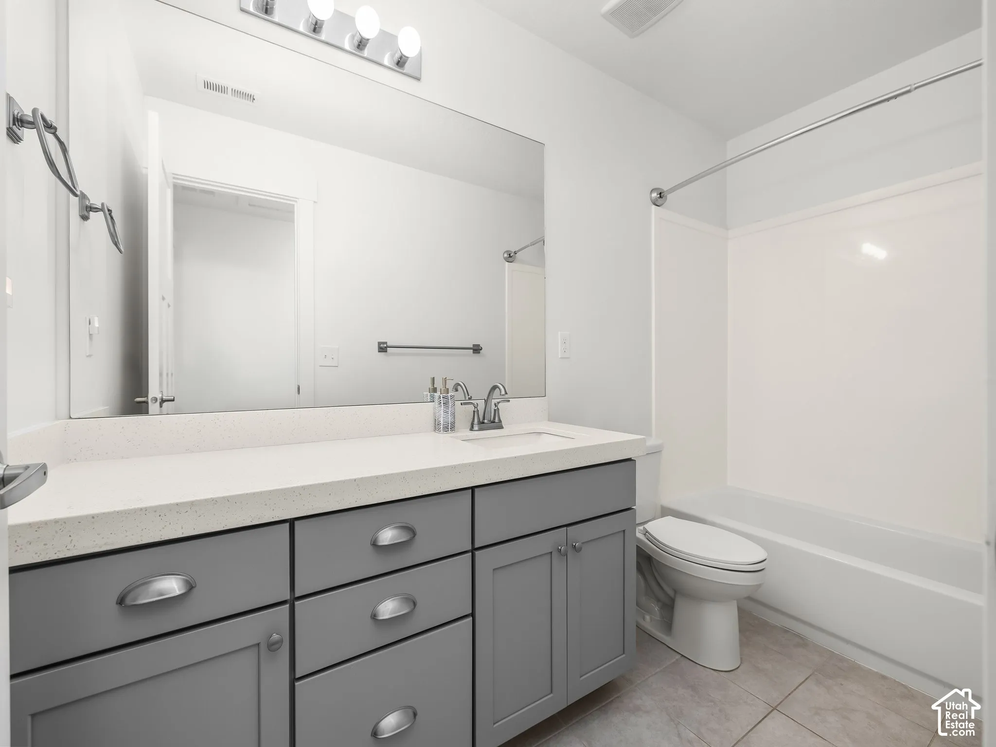 Bathroom featuring a grey shaker-style vanity, solid-surface countertop with an integrated sink, full-width vanity mirror, polished chrome hardware, and a tub-shower combination