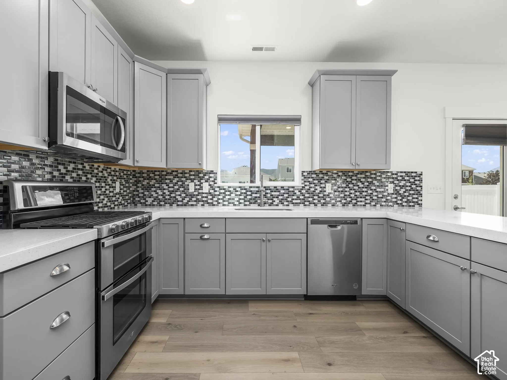 Kitchen featuring grey shaker-style cabinetry, white quartz countertops, stainless steel appliances, multi-toned glass tile backsplash, and wood-finish flooring