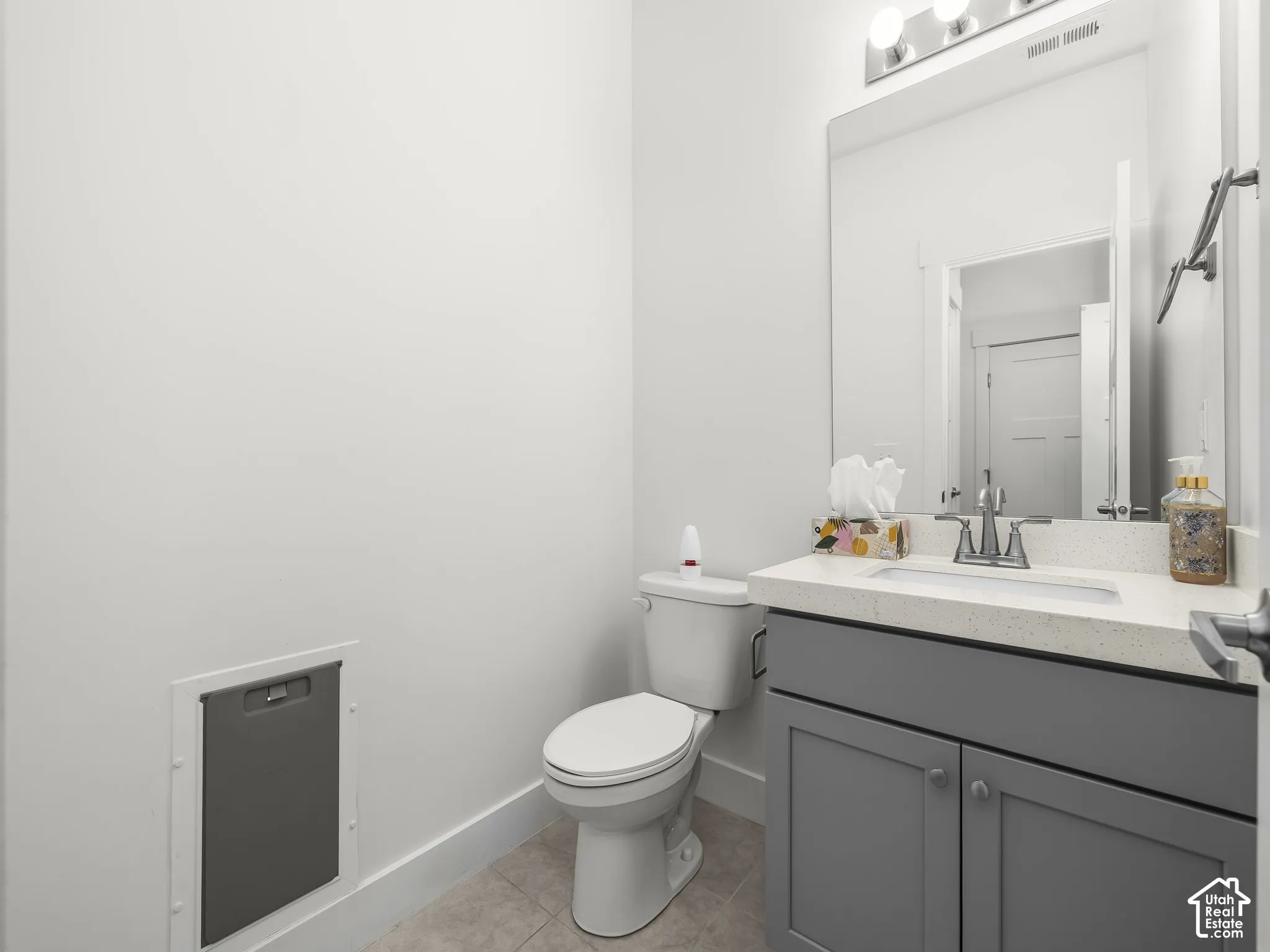 Grey shaker-style vanity with a light-toned solid surface countertop, rectangular undermount sink, three-light polished chrome fixture, and neutral tile flooring