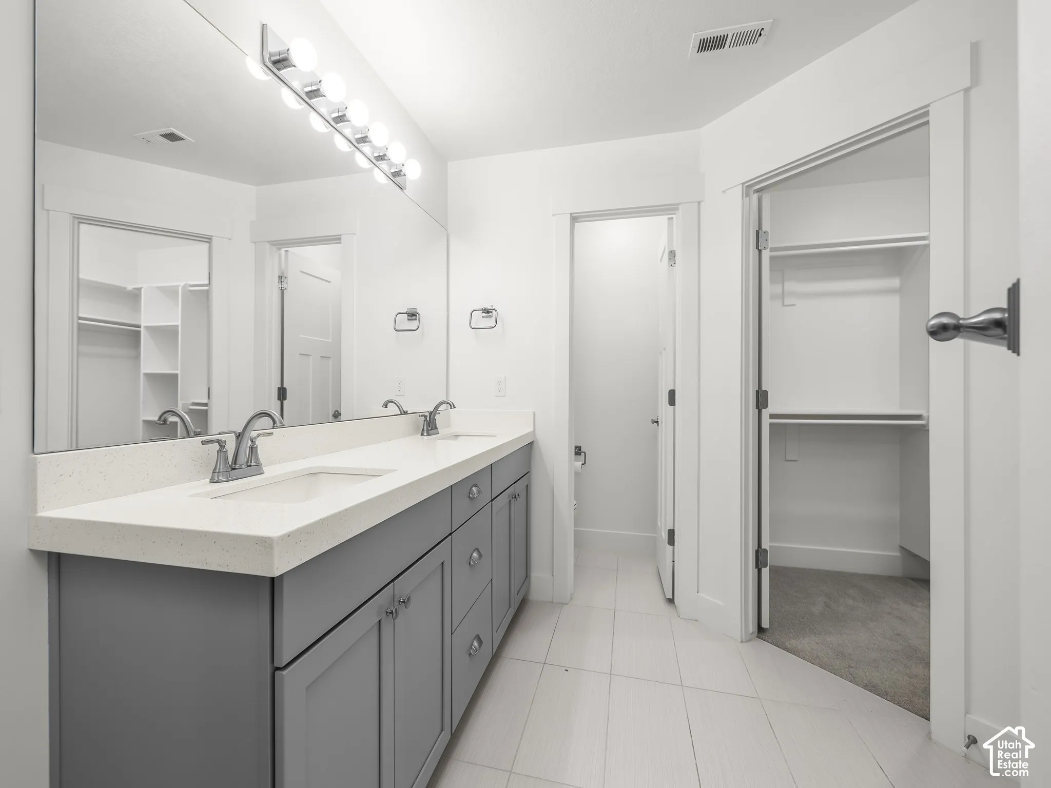 Dual-sink vanity with light-toned solid surface countertop, gray shaker-style cabinetry, full-width wall mirror, and integrated lighting