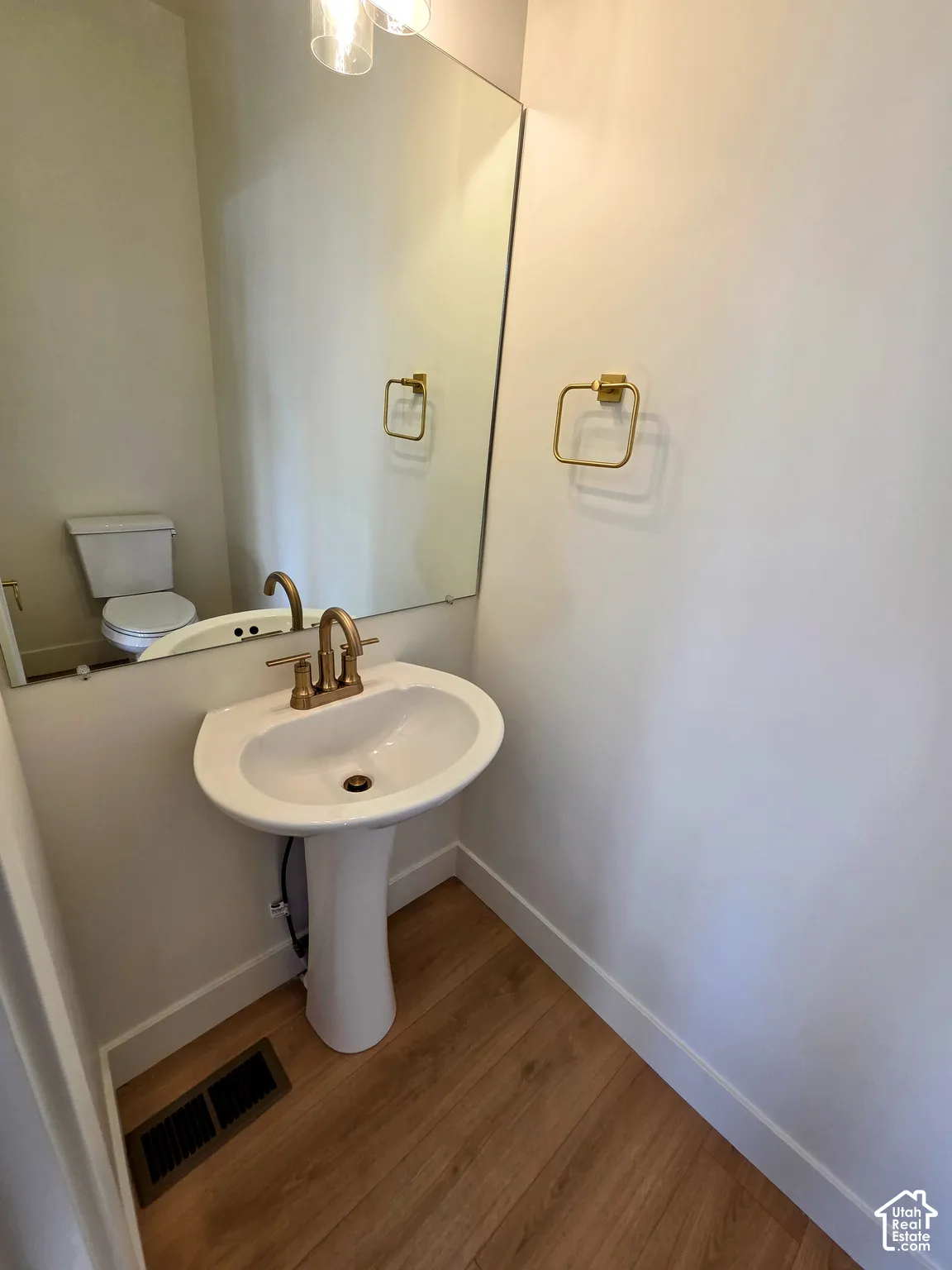 Pedestal sink with gold-tone faucet, wood-finish flooring, wall-mounted mirror, and gold-tone towel ring