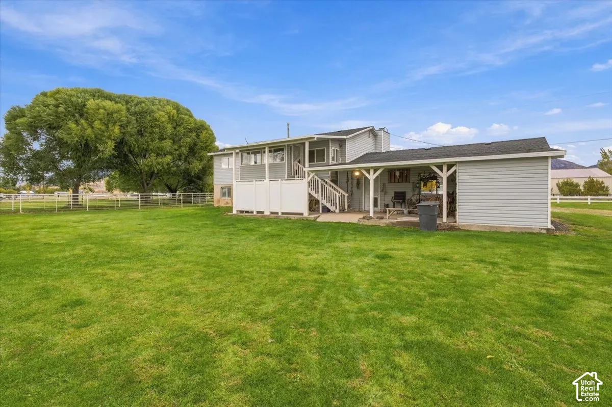 Expansive backyard featuring a manicured lawn, perimeter rail fencing, mature trees, and a covered patio with an attached carport
