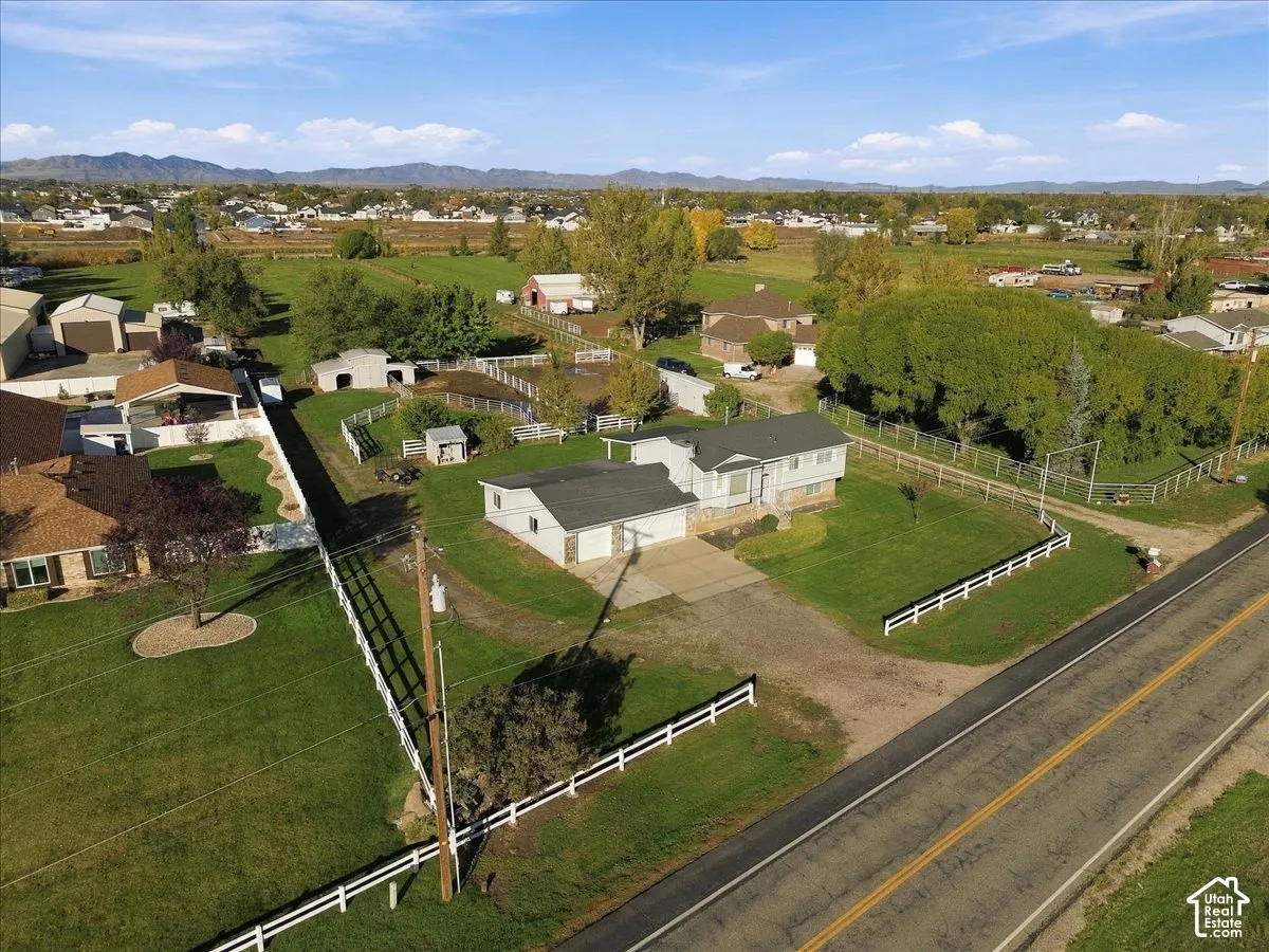 Aerial view of a multi-structure property featuring a residence with an attached garage, white perimeter fencing, mature trees, and mountainous terrain in the distance