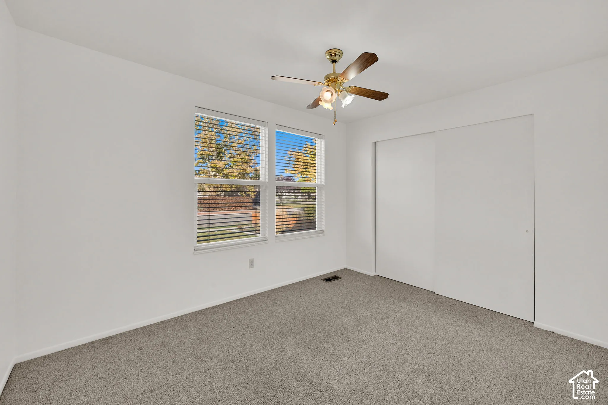 Neutral interior room featuring wall-to-wall carpeting, dual sliding closet doors, a brass-finish ceiling fan with integrated lighting, and two windows with horizontal blinds