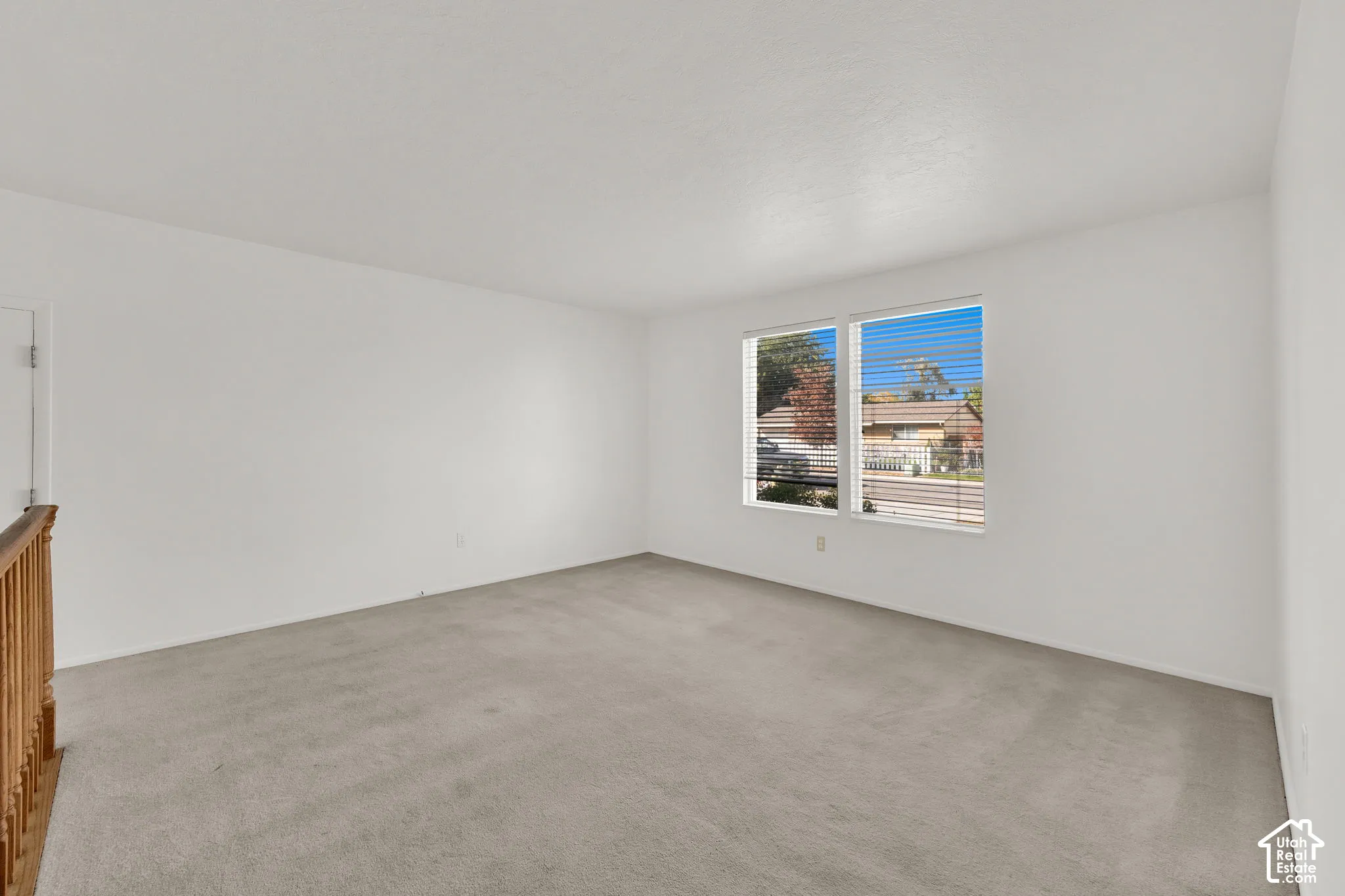 Neutral interior room featuring neutral-tone carpeting, smooth wall surfaces, and dual windows with horizontal blinds