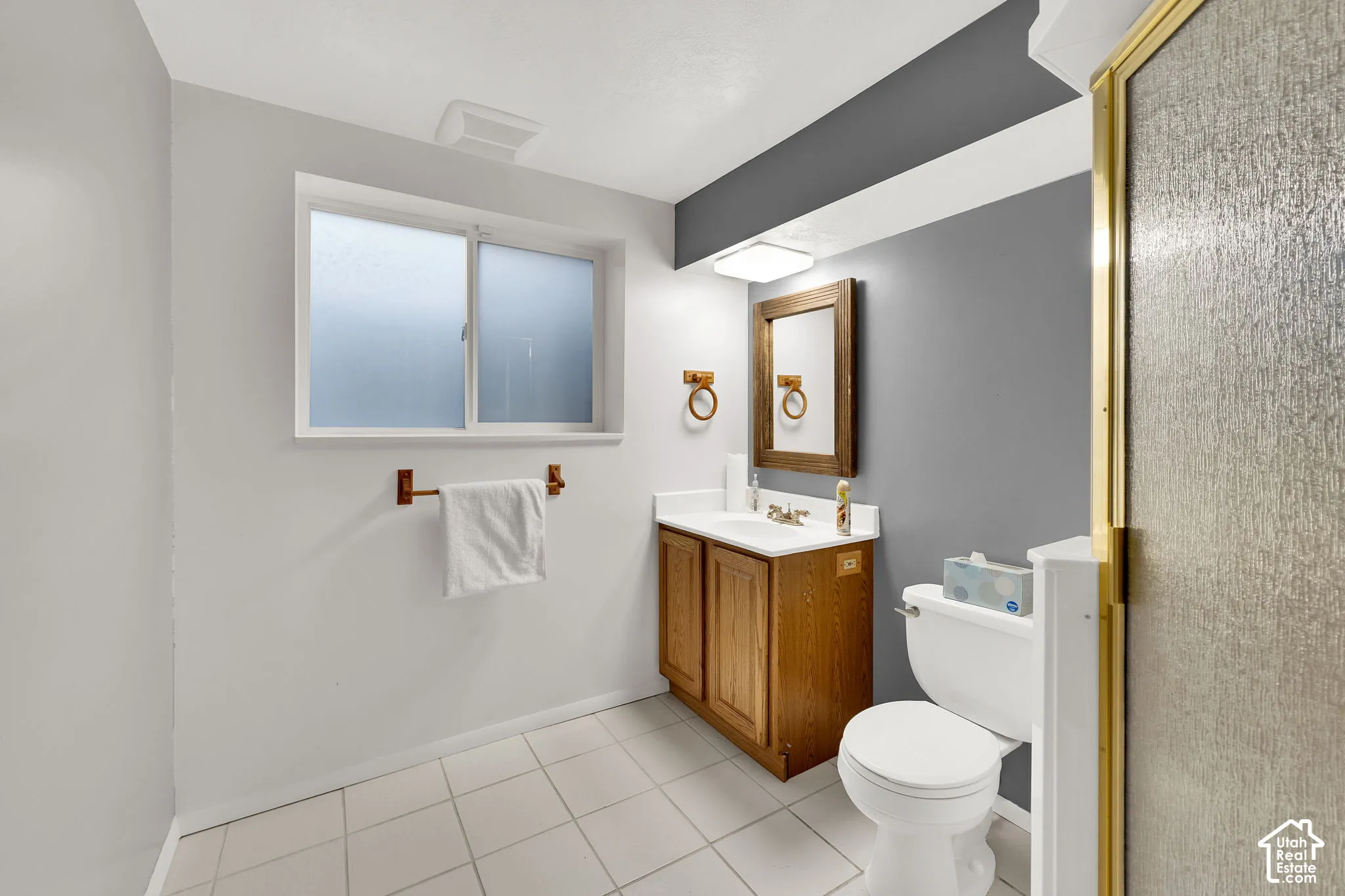 Bathroom featuring white tile flooring, wood cabinetry with a light-toned countertop, a sliding window with frosted glass, and a glass-enclosed shower with gold-toned frame