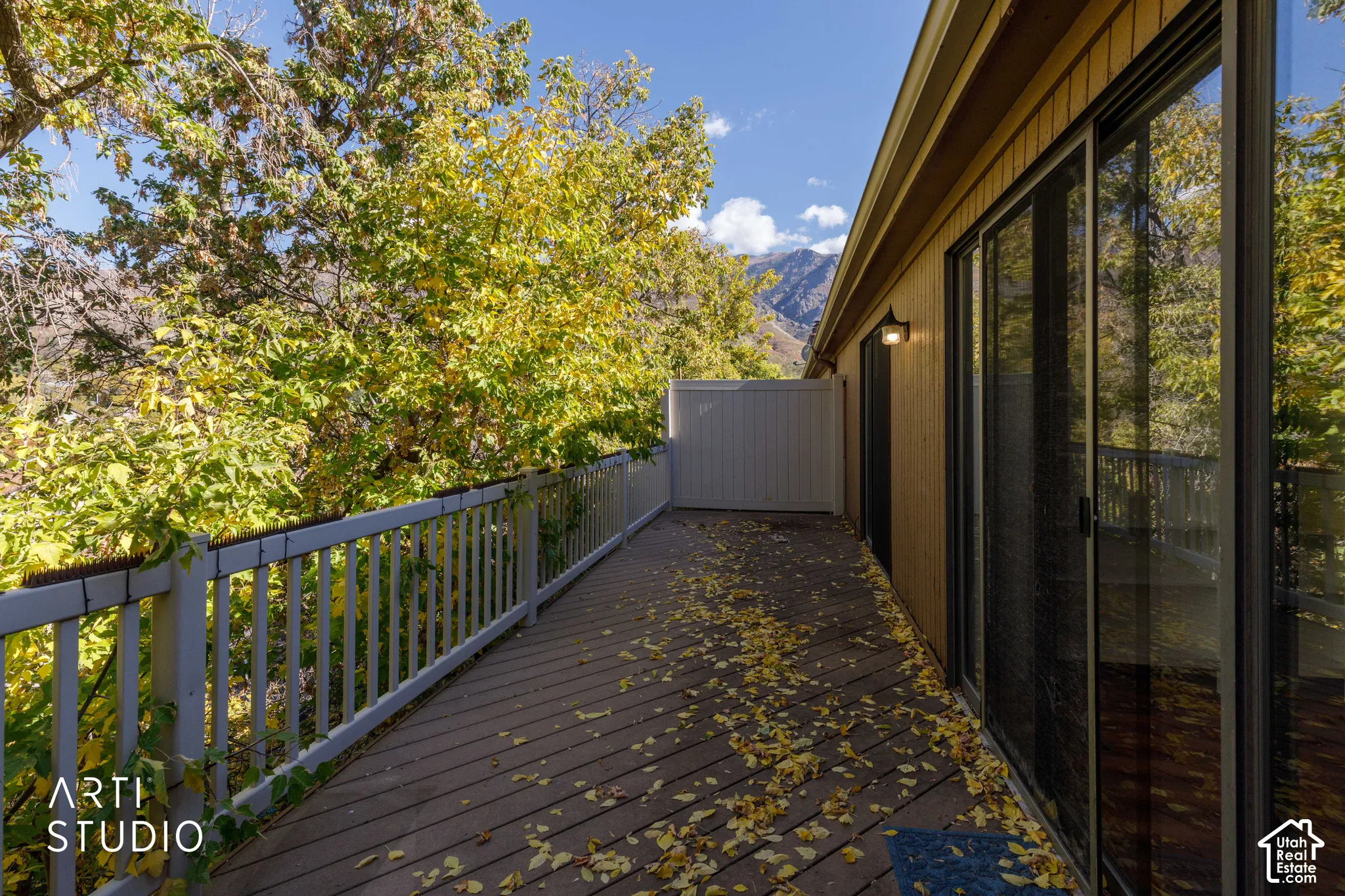 Exterior deck featuring wood-finish decking, white vertical balustrades, integrated wall-mounted exterior lighting, sliding glass door access, and mountain backdrop