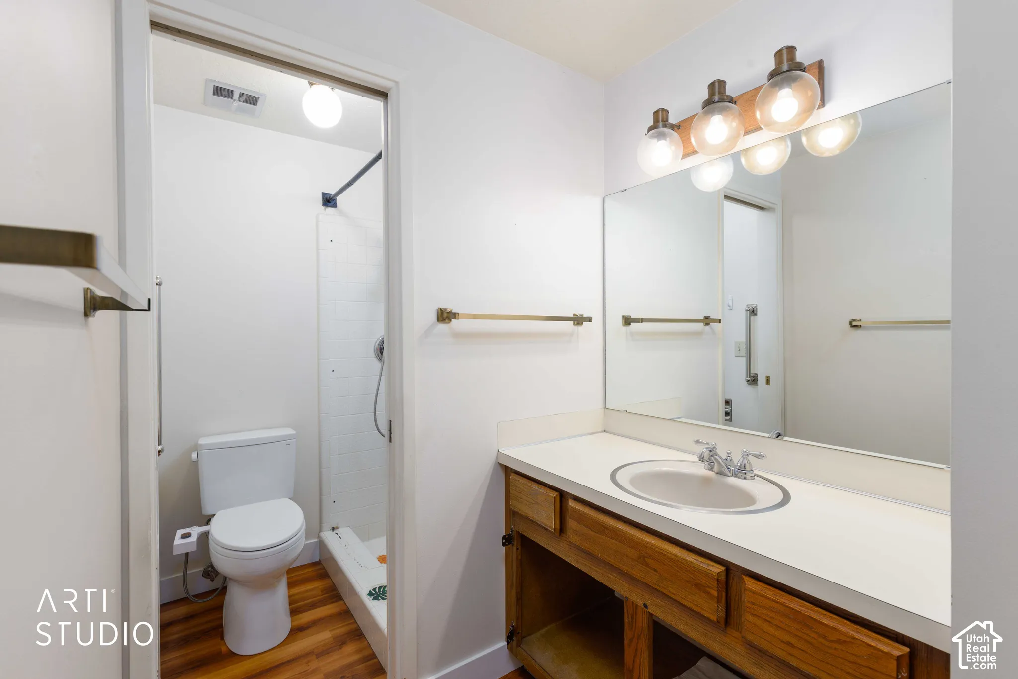 Bathroom featuring a wood-finish vanity with an integrated countertop sink, large wall-mounted mirror, multi-bulb overhead vanity lighting, and wood-finish flooring