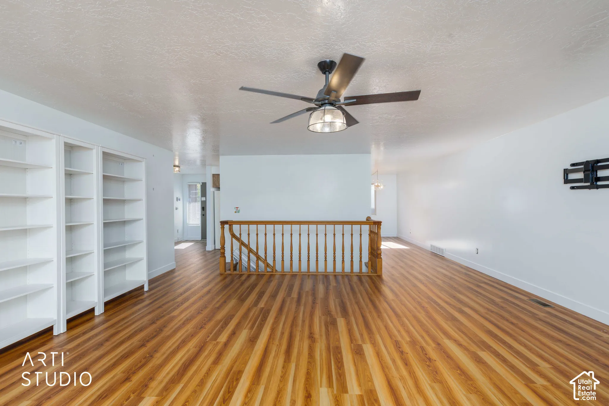 Open interior space featuring wood-finish flooring, built-in shelving, a ceiling fan with integrated lighting, and a wooden stair railing