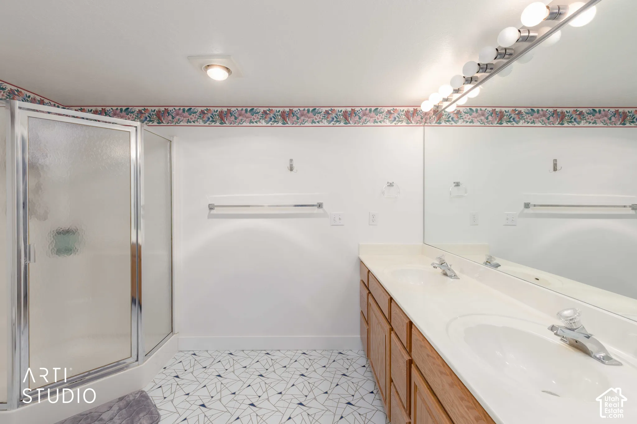 Bathroom featuring a corner shower enclosure with textured glass doors, a dual-sink vanity with wood-tone cabinetry, a full-width wall mirror, and geometric patterned flooring