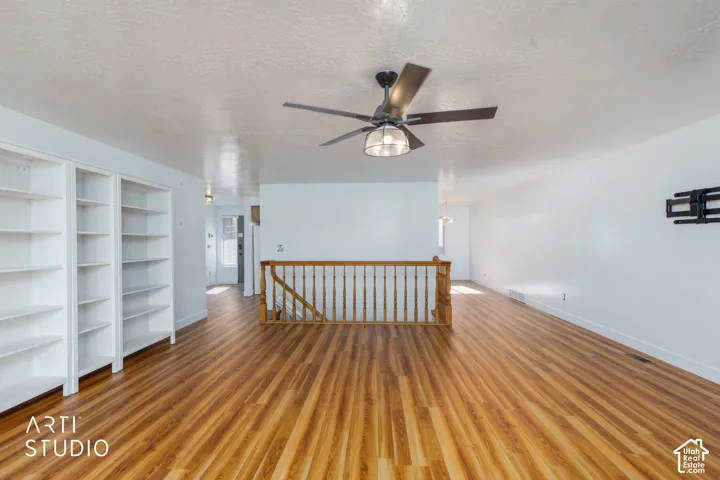 Open interior space featuring wood-finish flooring, built-in shelving, a ceiling fan with integrated lighting, and a wooden stair railing