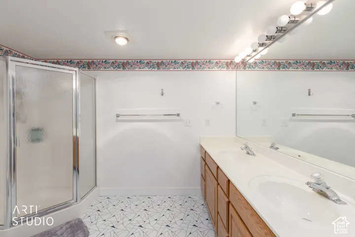 Bathroom featuring a corner shower enclosure with textured glass doors, a dual-sink vanity with wood-tone cabinetry, a full-width wall mirror, and geometric patterned flooring