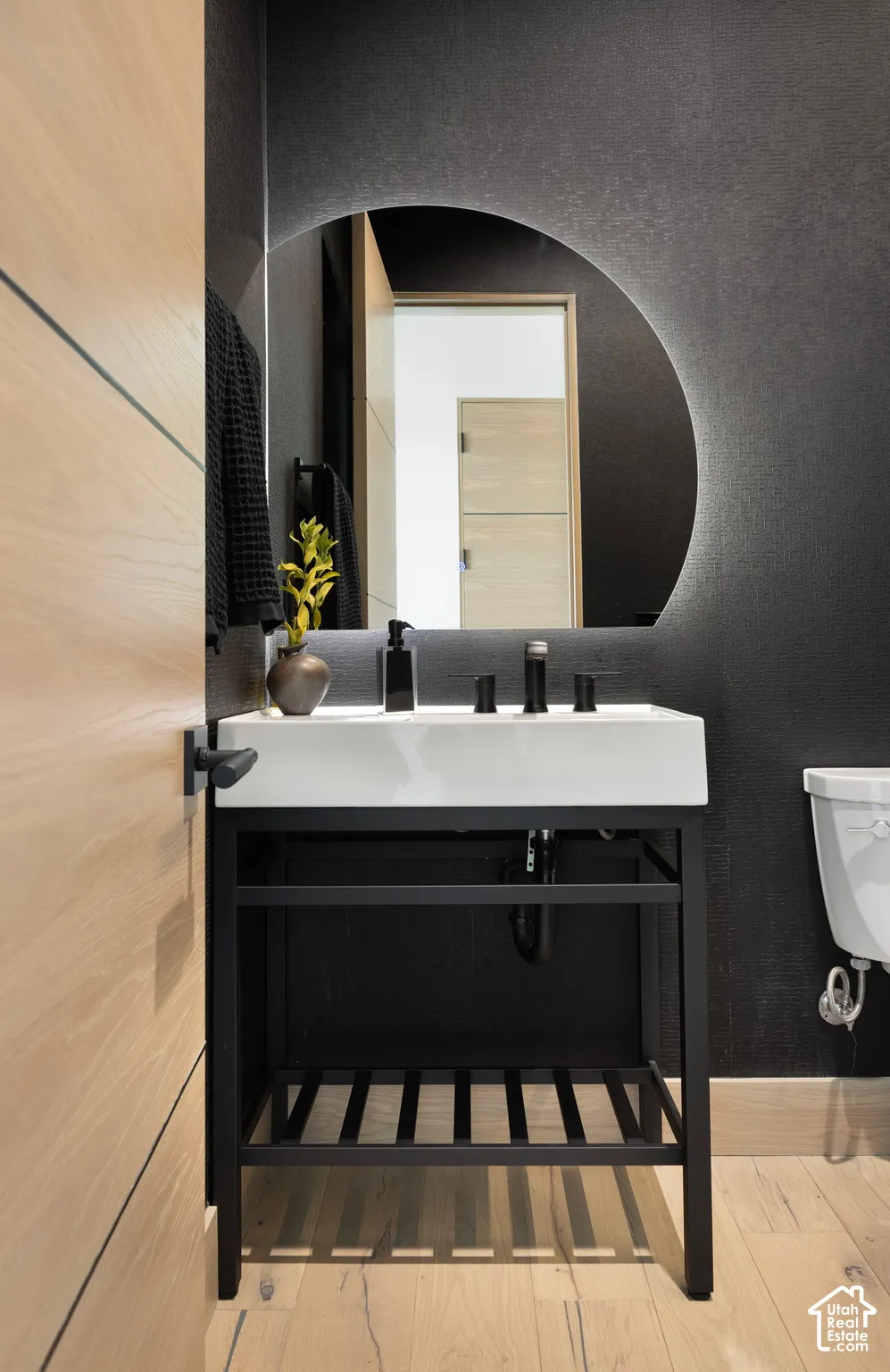 Modern vanity with a rectangular sink, black metal frame, integrated storage shelf, backlit circular mirror, and matte black widespread faucet
