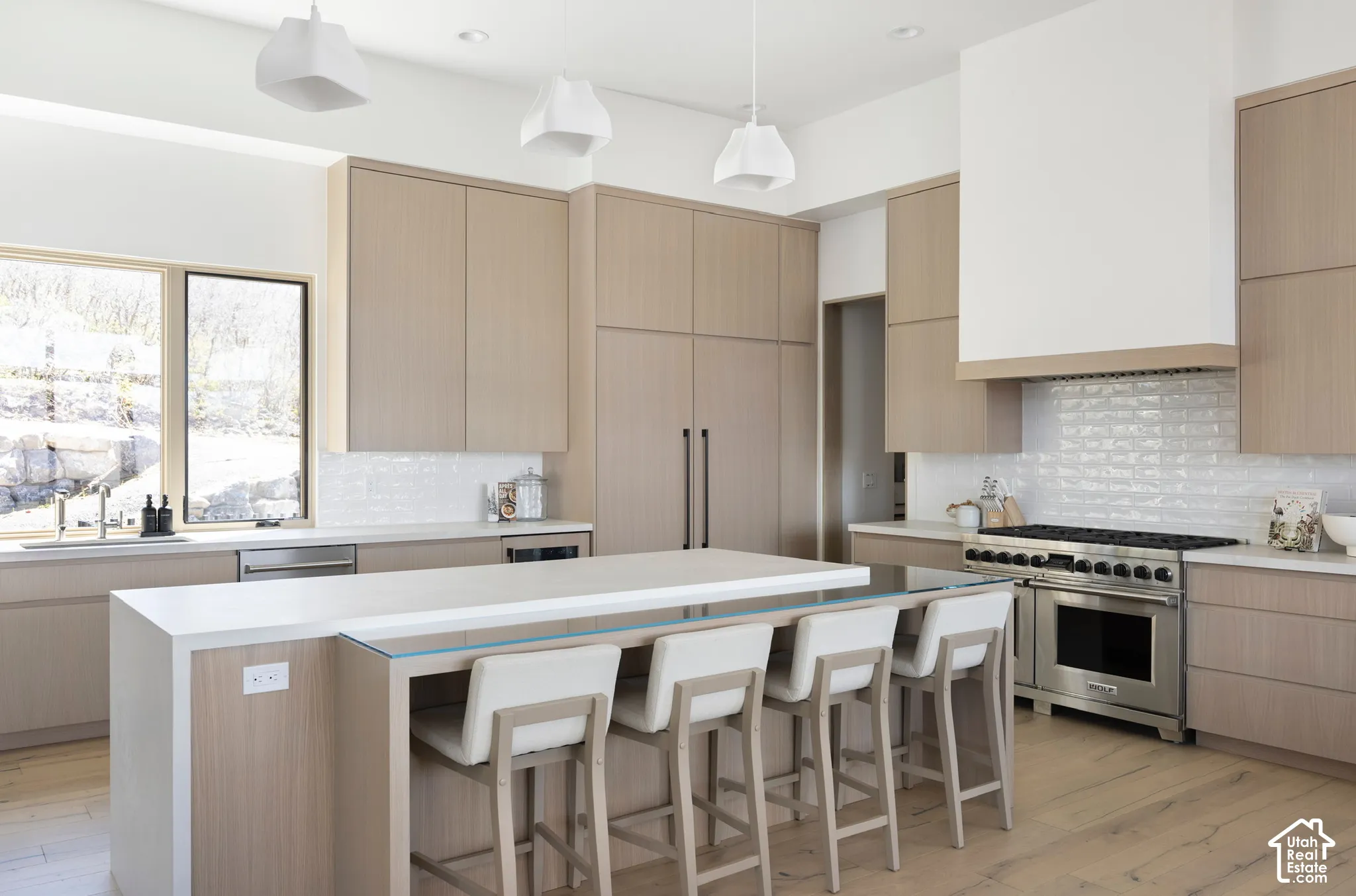 Kitchen featuring light wood-finish cabinetry, central island with an integrated glass-top counter, stainless steel professional-grade range, white subway tile backsplash, and three pendant lights