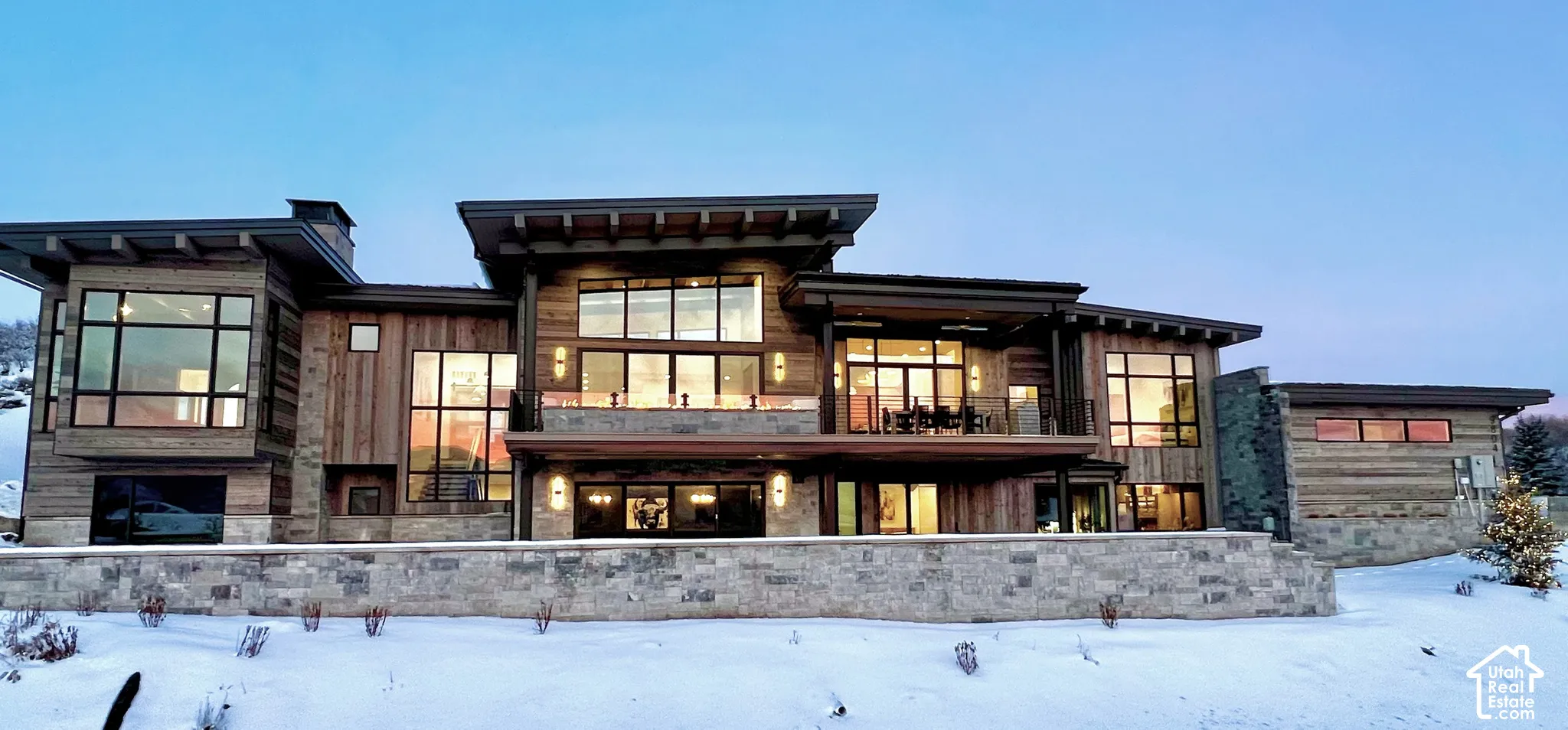 Modern mountain-style exterior featuring vertical wood siding, multi-level flat rooflines with deep eaves, expansive floor-to-ceiling windows, stacked stone masonry, and an elevated terrace