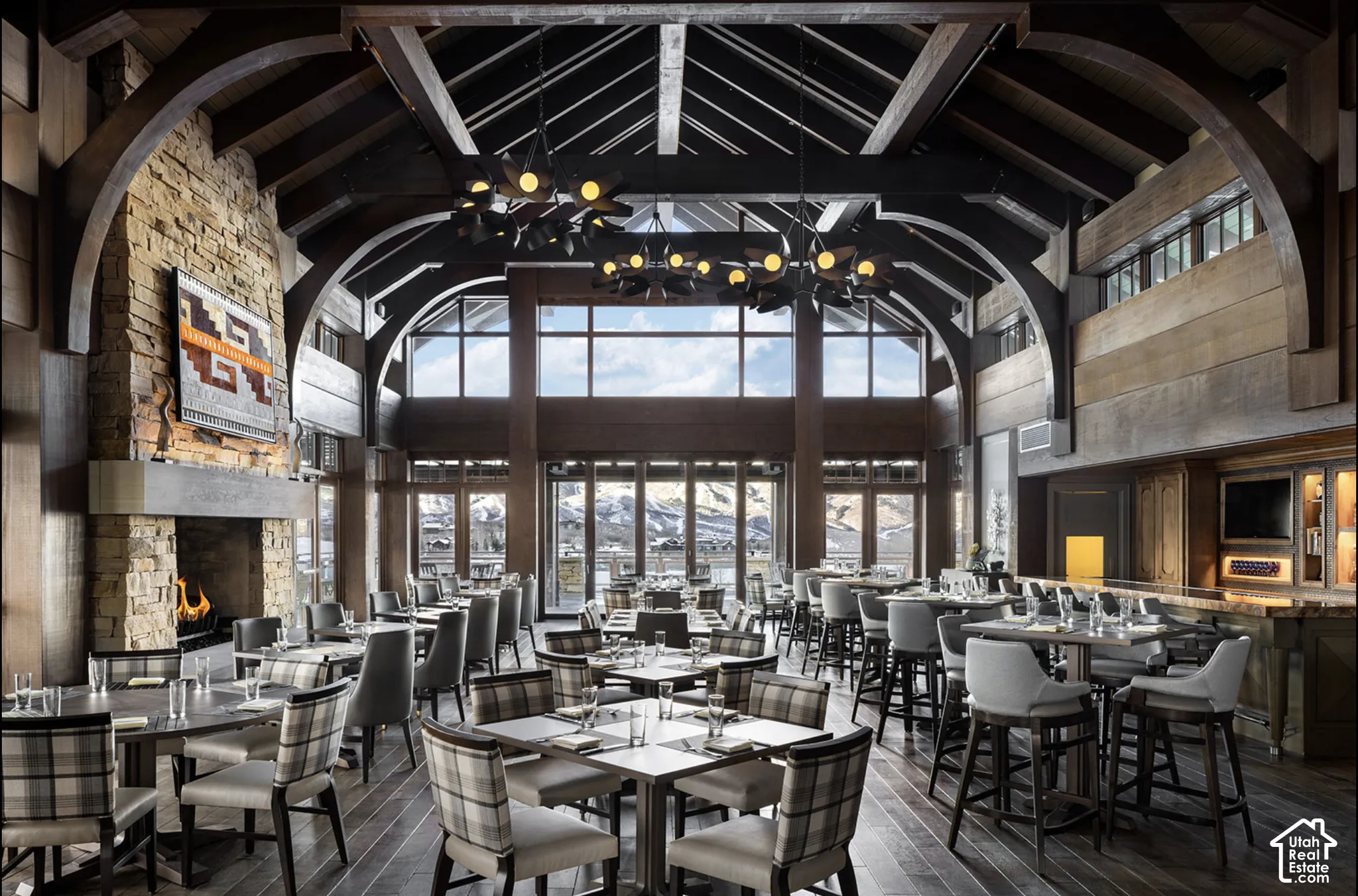 Vaulted ceiling with exposed timber trusses, floor-to-ceiling windows, stone-surround fireplace, wood-finish flooring, and integrated bar cabinetry