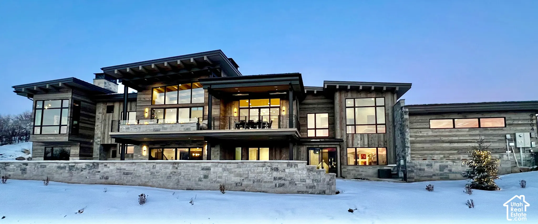 Contemporary exterior featuring wood-plank siding, natural stone accents, expansive floor-to-ceiling windows, deep roof overhangs with exposed rafters, and a tiered stone retaining wall