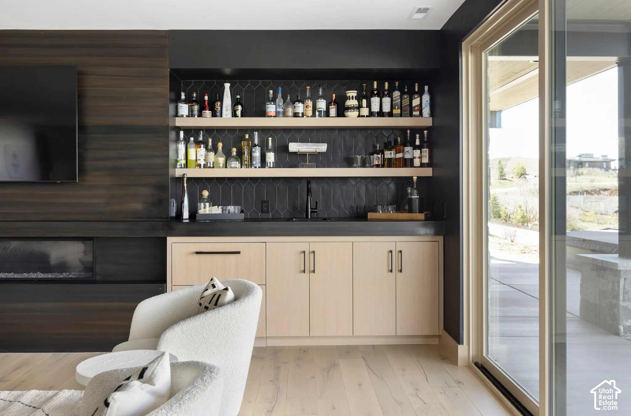 Built-in wet bar featuring light wood cabinetry, stone countertops, matte black plumbing fixtures, floating shelves, and dark vertical tile backsplash