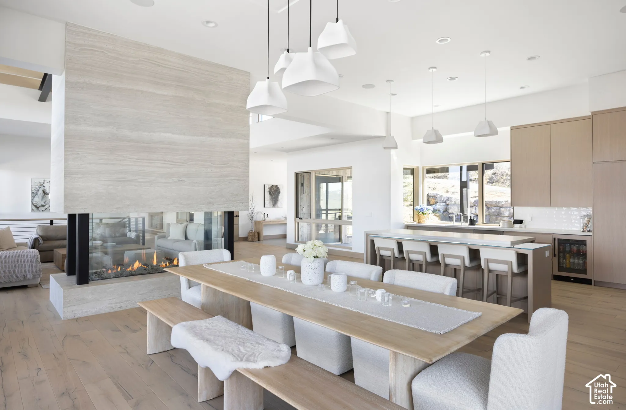 Open-concept space featuring wood-finish flooring, a floor-to-ceiling stone-clad double-sided fireplace, minimalist pendant lighting, and built-in cabinetry with integrated appliances