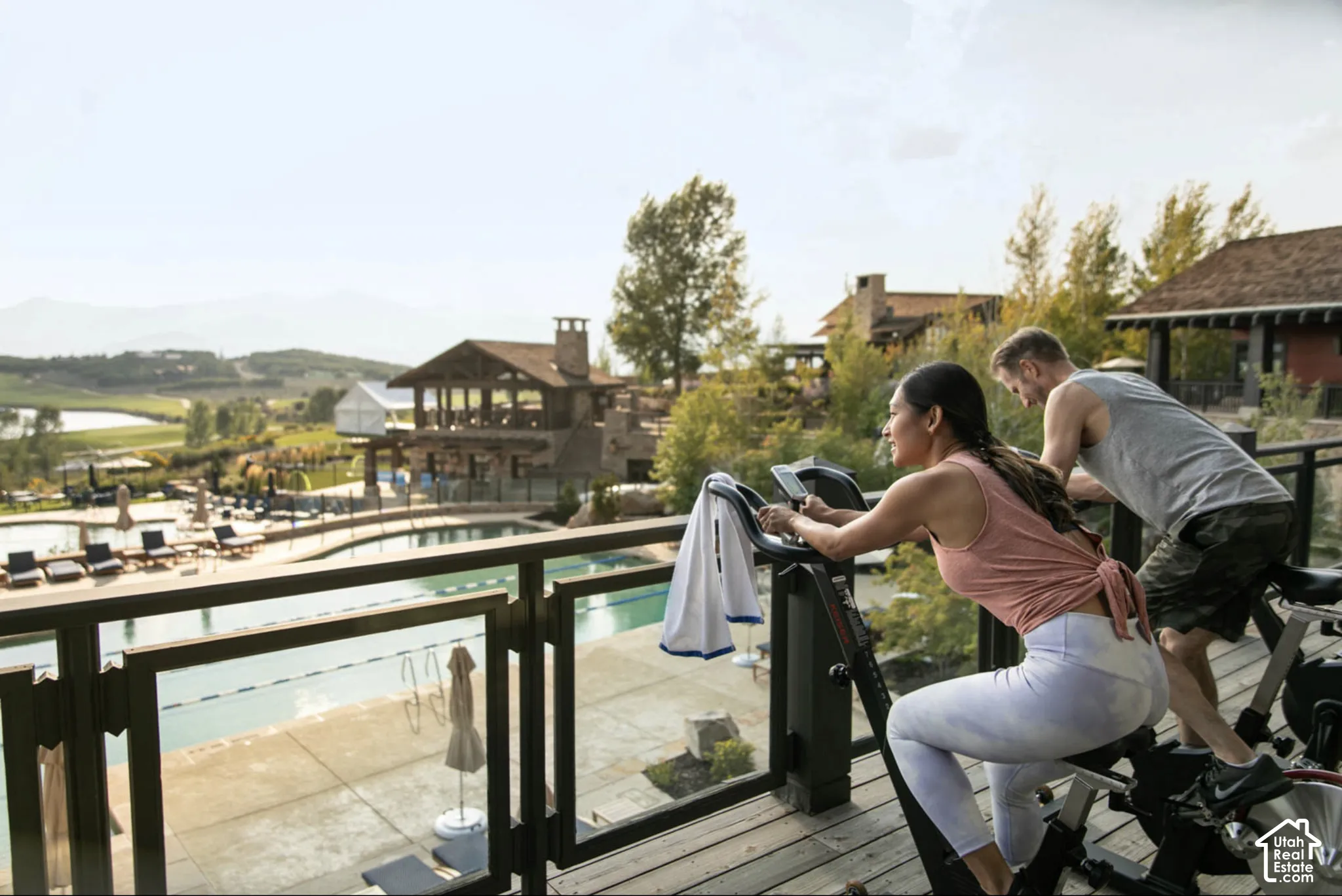 Elevated wooden deck featuring a glass and metal railing, overlooking a large swimming pool, stone patio, and distant mountain scenery