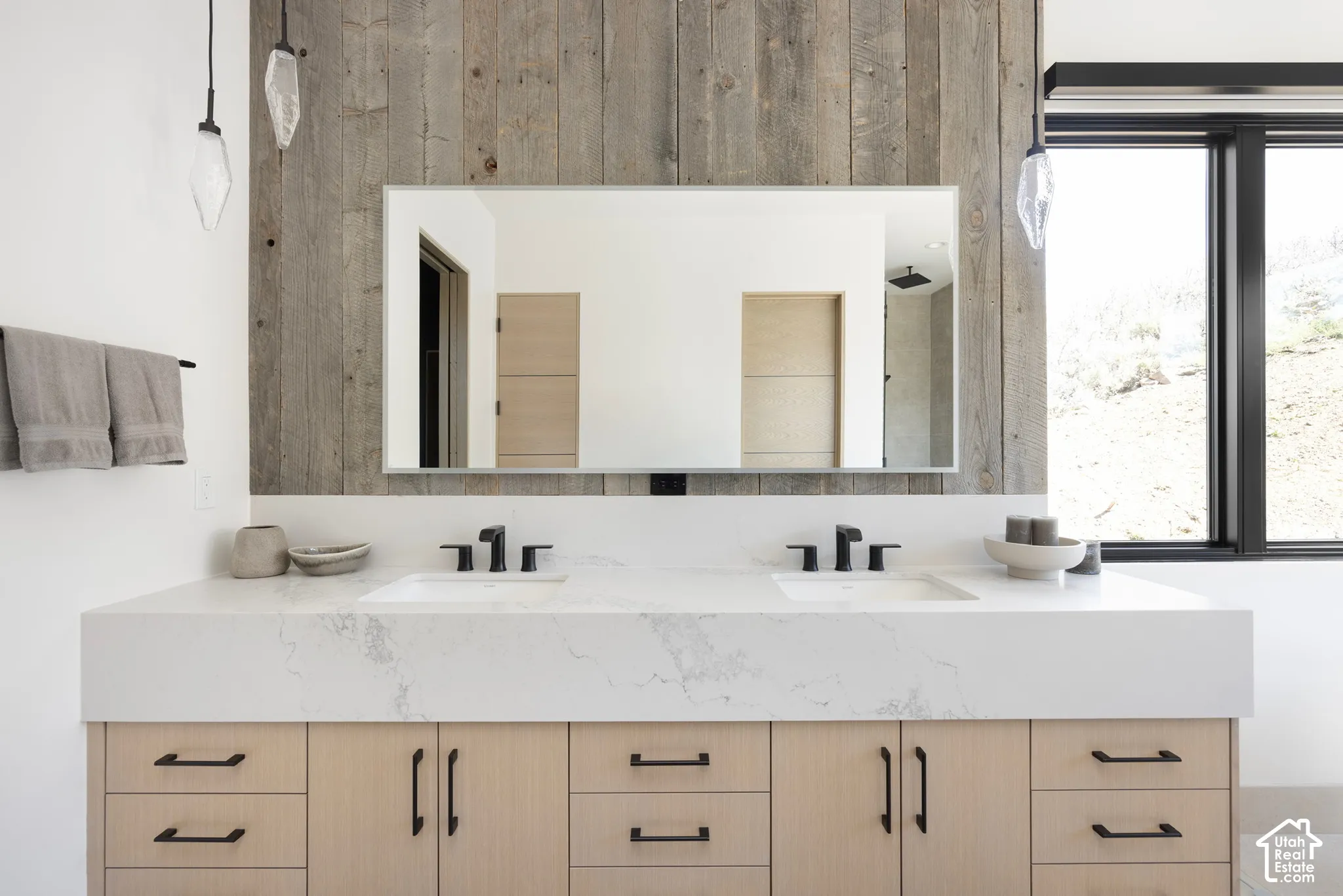 Dual-vanity featuring light-tone cabinetry, stone-surface countertop with integrated sinks, matte black fixtures, and a reclaimed wood-panel accent wall