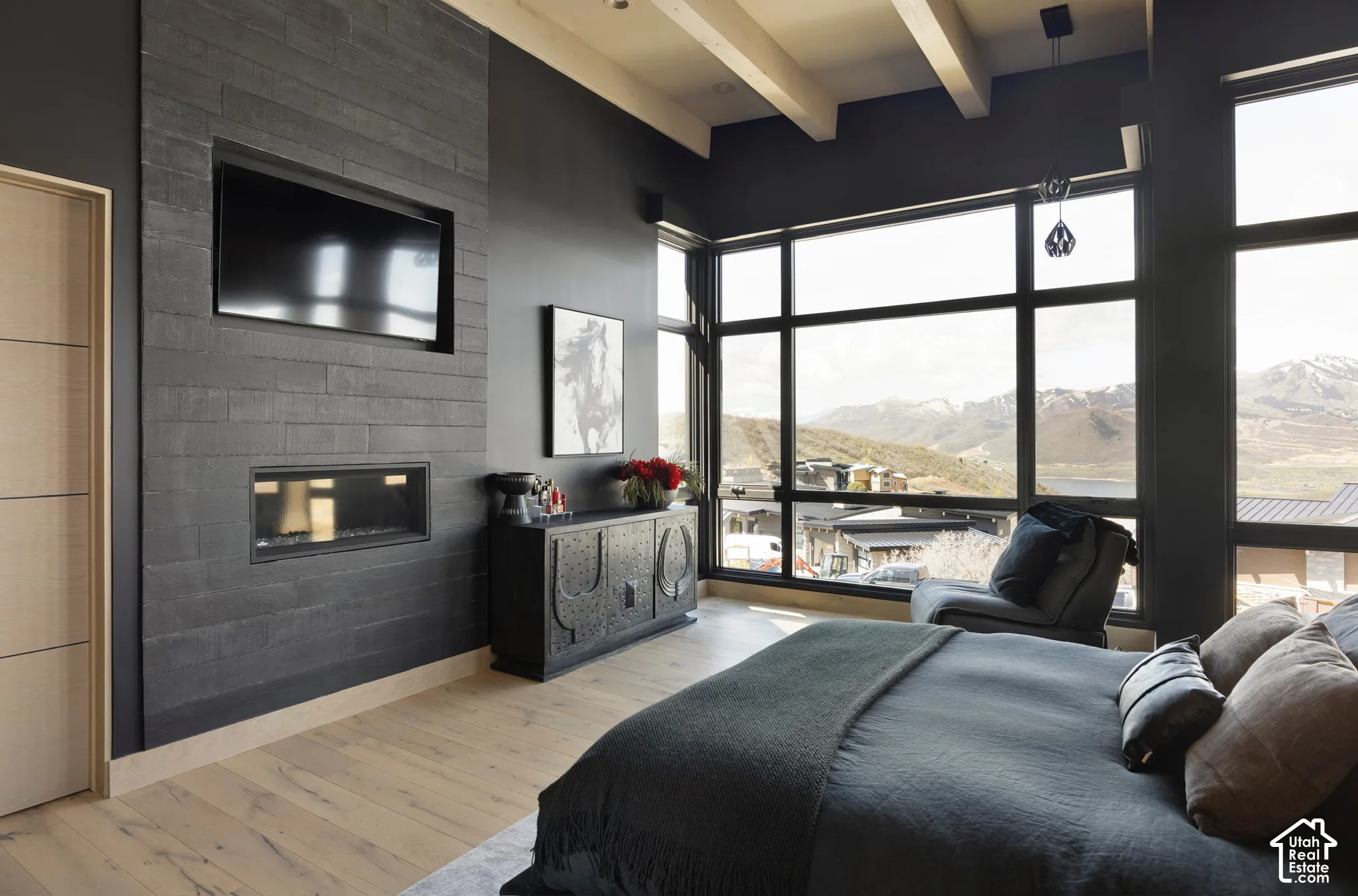 Full-height stone-clad fireplace featuring an integrated television niche, expansive floor-to-ceiling windows, exposed structural ceiling beams, and wood-finish flooring