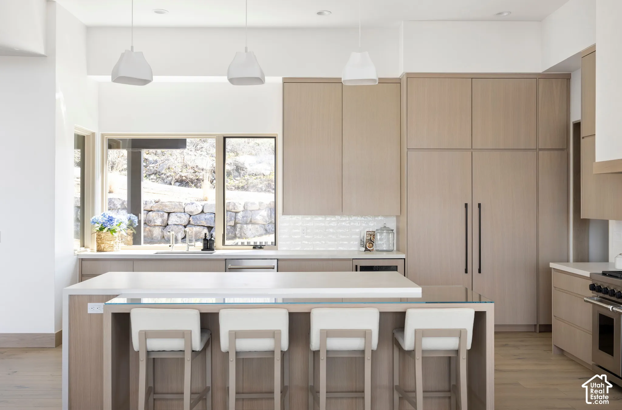 Kitchen featuring light wood-finish cabinetry, a waterfall-edge island, textured tile backsplash, modern pendant lighting, and stainless steel integrated appliances