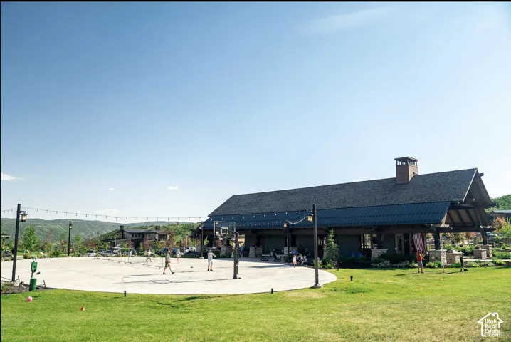 Outdoor recreation area featuring a paved sports court with a basketball hoop, covered pavilion with stone pillar supports, dark metal roofing, and perimeter string lighting