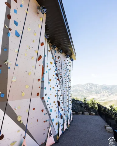 Custom outdoor rock climbing wall with integrated climbing holds, multiple rope anchors, high-intensity mounted floodlights, and expansive mountain terrain backdrop