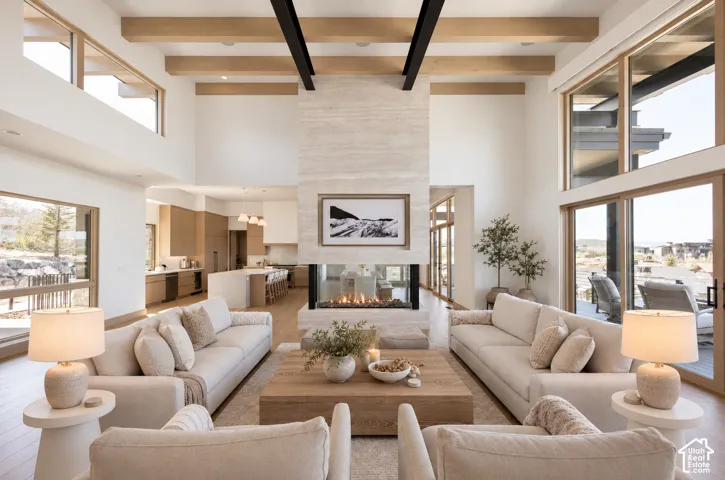 Double-height ceiling with structural wood-finish beams, stone-clad dual-sided fireplace, expansive windows, and wide-plank wood-finish flooring
