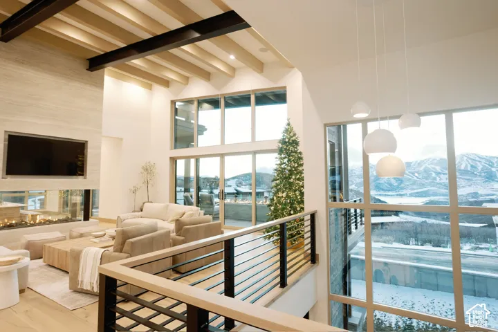 Open-concept interior featuring exposed structural beams, floor-to-ceiling windows, mountain scenery, custom stone-surround fireplace, and a metal-rail staircase