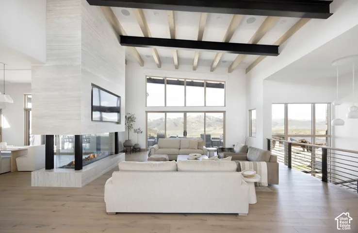 Double-height great room featuring exposed structural steel beams, light wood ceiling beams, floor-to-ceiling stone fireplace with multi-sided glass firebox, wood-finish flooring, and expansive windows