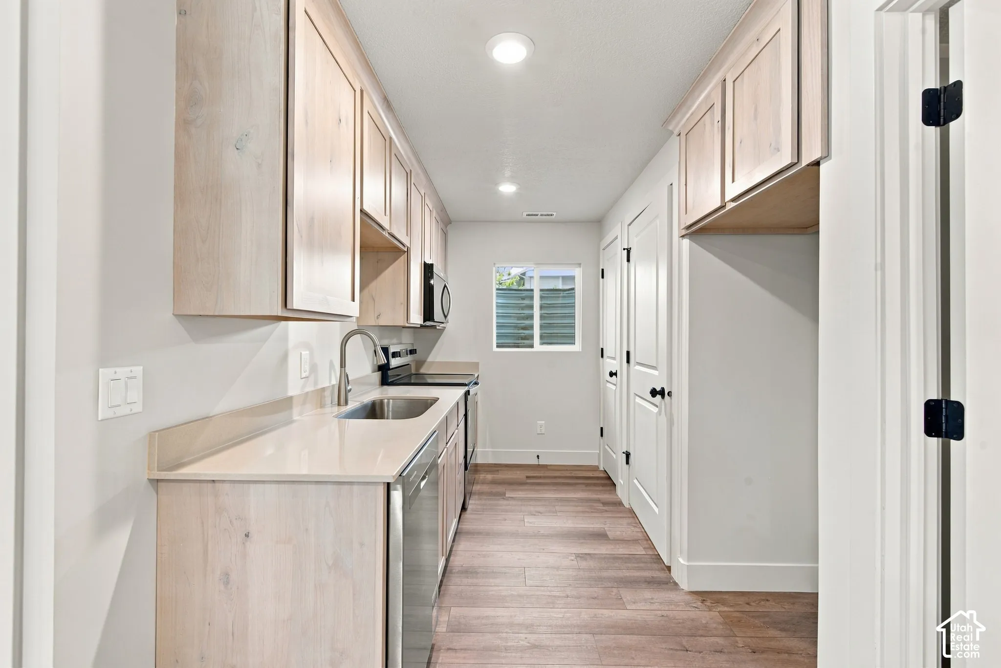 Galley kitchen featuring light-toned wood-finish cabinetry, solid surface countertops, stainless steel appliances, and wood-finish flooring