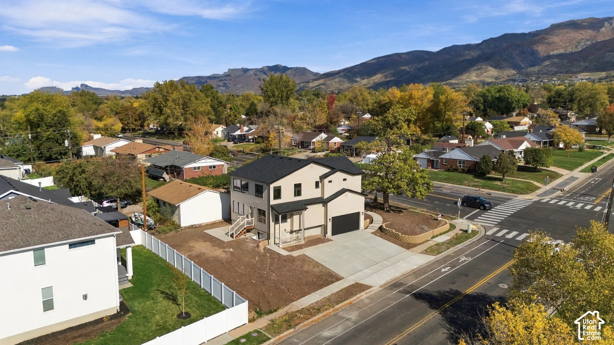 Two-story residence featuring a dark shingle roof, concrete driveway, attached garage, and a white vinyl fence, set against a backdrop of mountain scenery