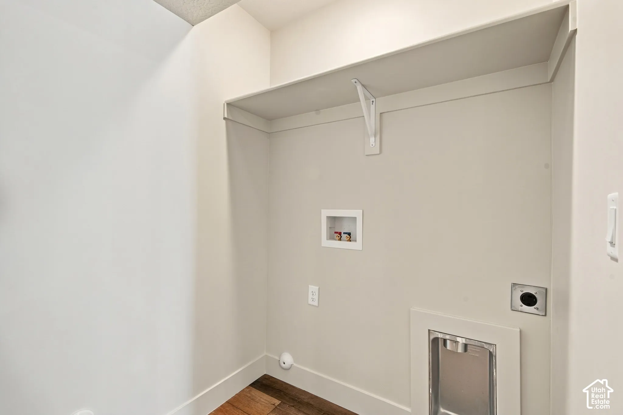 Utility area featuring wood-finish flooring, a white overhead storage shelf with bracket support, and recessed water and dryer hookups