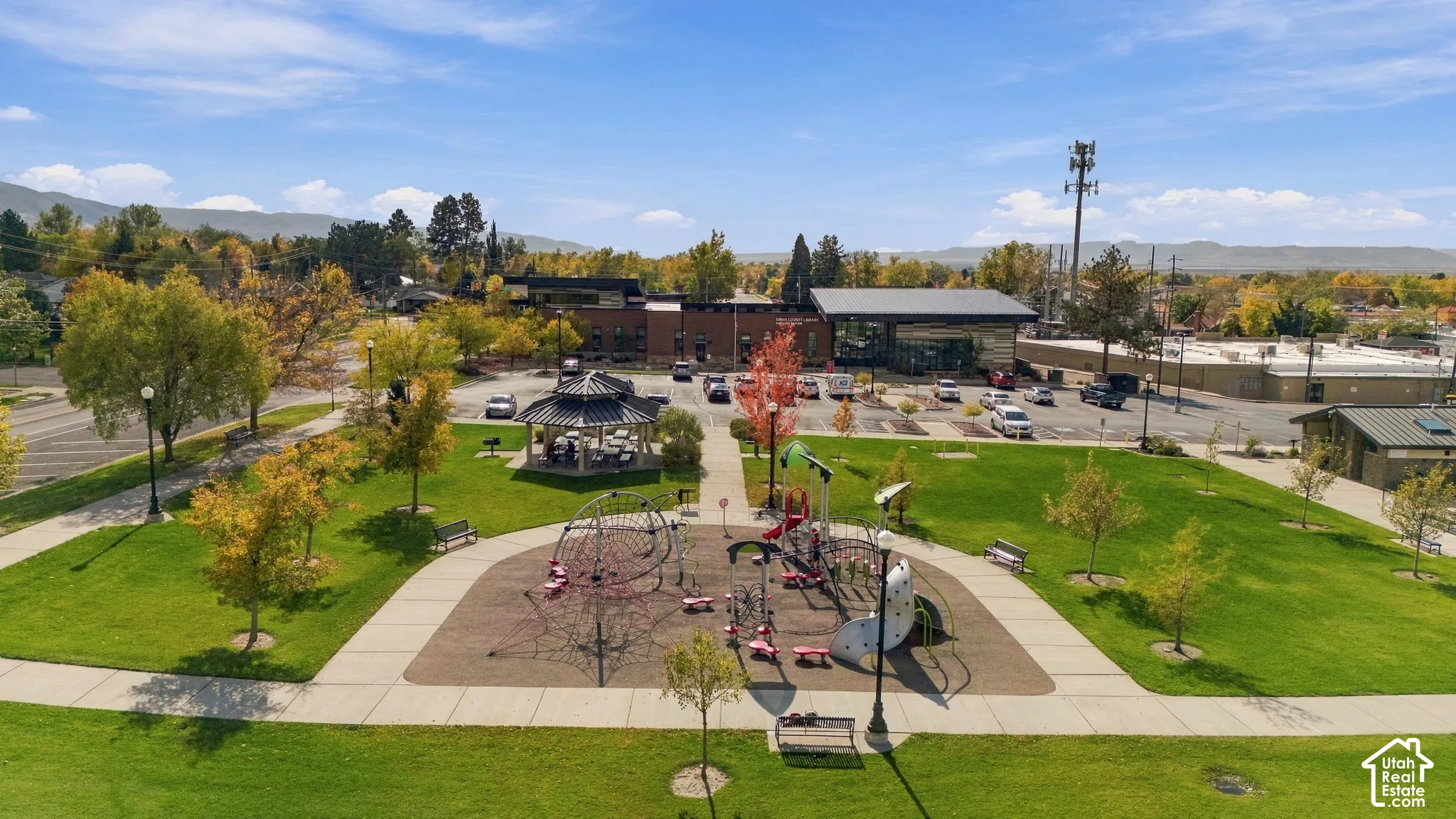 Public park featuring a manicured lawn, paved walkways, playground structures, a covered gazebo, and mature trees