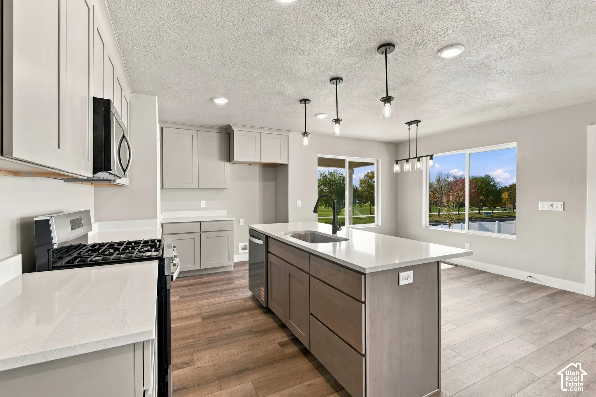 Kitchen featuring light gray cabinetry, solid-surface countertops, wood-finish flooring, a center island with an undermount sink and matte black faucet, and pendant lighting
