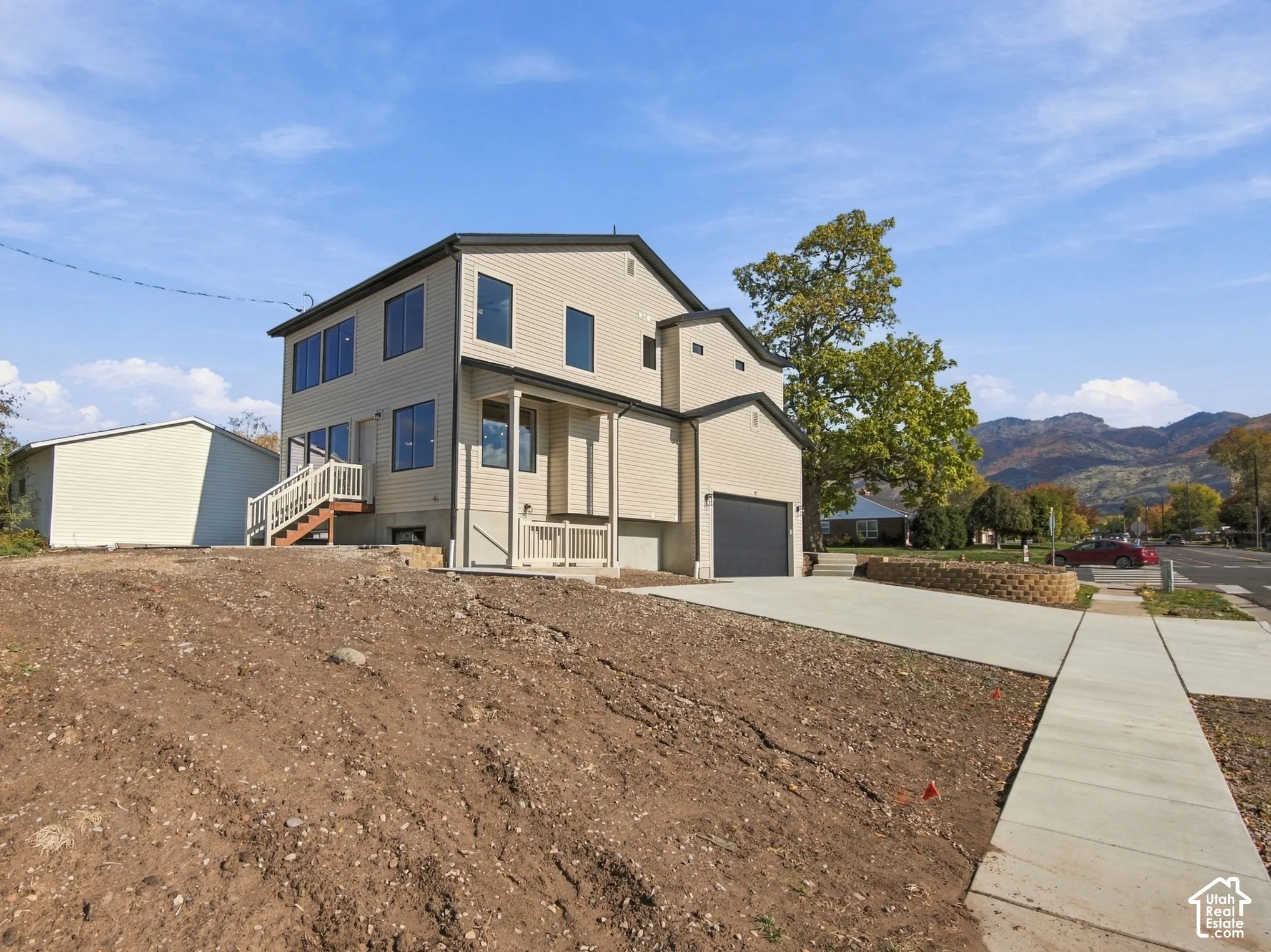 Two-story exterior featuring tan horizontal siding, an attached garage, a concrete driveway, a raised wood deck, and mountain backdrop