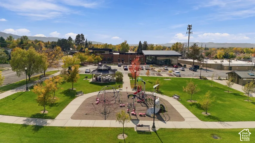Public park featuring a manicured lawn, paved walkways, playground structures, a covered gazebo, and mature trees