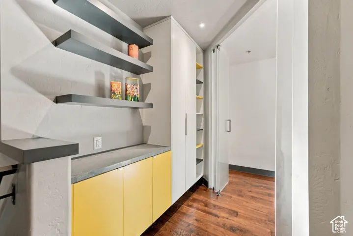 Built-in cabinetry with custom yellow base units, stone-finish countertops, floating shelving, and a full-height glass panel door featuring wood-finish flooring